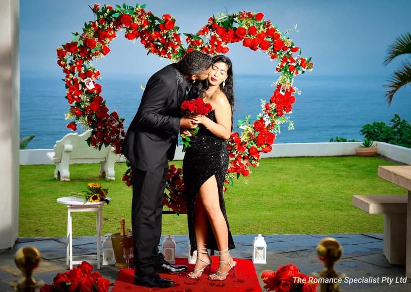 Violan kisses Bianca's cheek beneath a heart-shaped arch of red roses overlooking the sea, a bouquet held between them